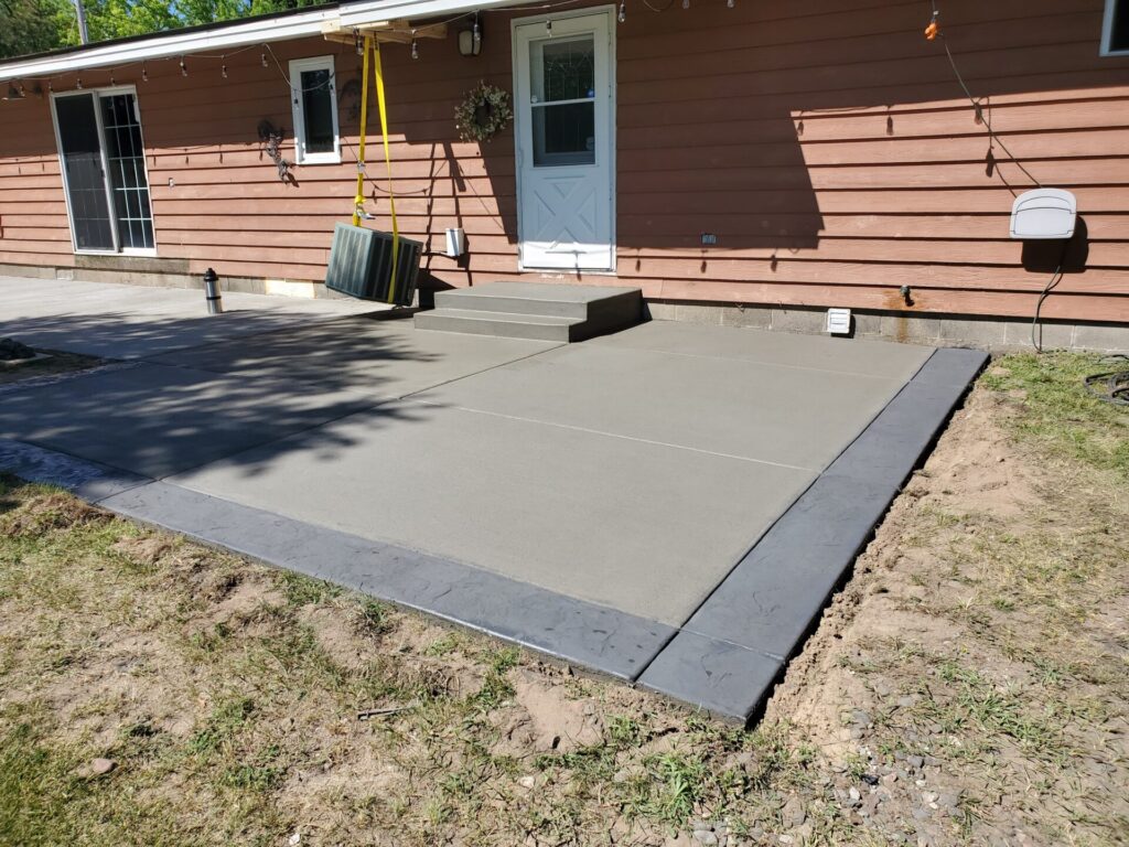 Freshly poured concrete patio with a textured border next to a house with a red exterior and white door.