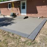 Freshly poured concrete patio with a textured border next to a house with a red exterior and white door.