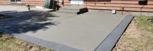 Freshly poured concrete patio with a textured border next to a house with a red exterior and white door.