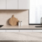 White stone product display table located in stylish blurry kitchen with beige walls, white cabinets and window. 3d rendering