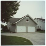 A two-car garage attached to a gray house, with a paved driveway and a tree partially visible on the left.
