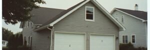 A two-car garage attached to a gray house, with a paved driveway and a tree partially visible on the left.
