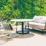 Outdoor patio with a round table, two rocking chairs, and a cushioned sofa set against a green leafy background.