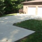 Freshly poured concrete driveway next to a patch of green grass in front of a brick house with two garage doors.