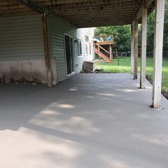 Concrete patio under a covered deck at the back of a house, with a grassy yard and trees in the background.
