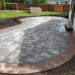 A newly installed stamped concrete patio with a brown border beside a white house in a backyard.