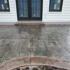 Stamped concrete patio in a rectangular pattern outside a house with white siding and black double doors.