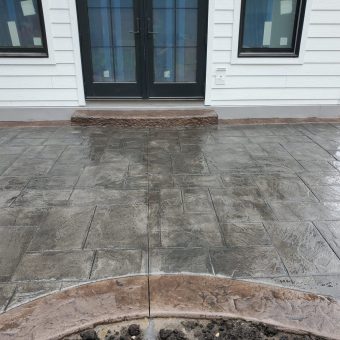 Stamped concrete patio in a rectangular pattern outside a house with white siding and black double doors.