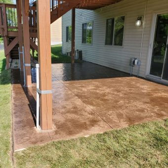 Concrete patio beneath a wooden deck attached to a house, with grass bordering the patio area.