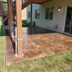 Concrete patio beneath a wooden deck attached to a house, with grass bordering the patio area.