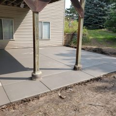 Freshly poured and smoothed concrete patio under a covered deck next to a house, with surrounding dirt and grass.