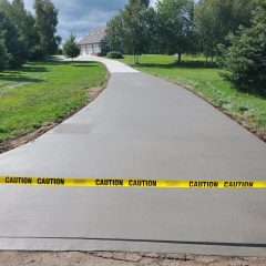 Freshly poured concrete driveway blocked off with caution tape, surrounded by grass and trees, house in background.