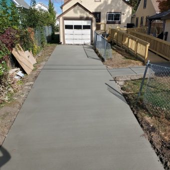 Freshly poured concrete driveway leads to a detached garage, with chain-link fences on both sides and a small sidewalk.