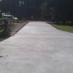 A newly poured concrete driveway extends toward a tree line, with caution tape visible in the background.