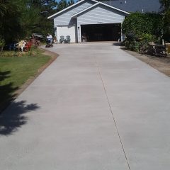 A long concrete driveway leads to a white house with an open garage and trees in the background.