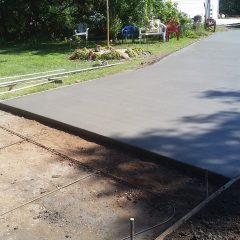 Freshly poured concrete driveway next to a yard with chairs, an American flag, and a metal garage in the background.