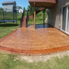 A stamped concrete patio with a decorative border next to a house, beneath a raised deck with stairs, in a backyard.