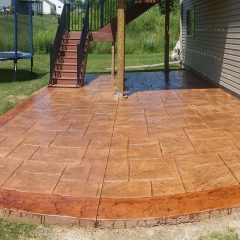 A stamped concrete patio with a patterned surface next to a house, with stairs leading up and a trampoline in the yard.