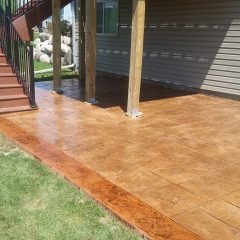 Stamped concrete patio under a deck with wooden support posts, next to a grassy lawn and house siding.