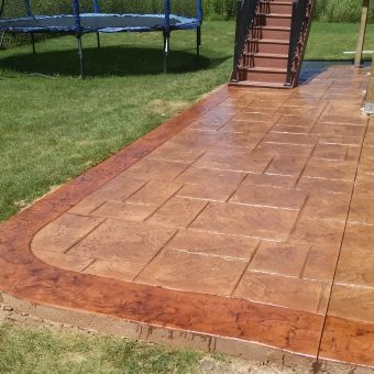 Stamped concrete patio with a geometric pattern beside grass, near a stairway and a trampoline in a backyard.
