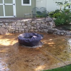 A covered fire pit sits on a stamped concrete patio in front of a stone wall and green shed in a backyard.