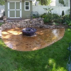 Backyard with a stamped concrete patio, a covered fire pit, stone wall, garden beds, and a small shed in the background.