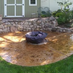 Stamped concrete patio with a covered fire pit, stone retaining wall, and garden beside a shed.
