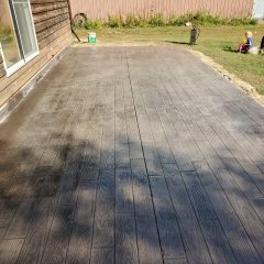 A wooden textured patio beside a house, with a lawn and a metal building in the background.