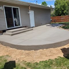 Freshly poured concrete patio with curved edge outside a beige house, bordered by dirt and grassy lawn.