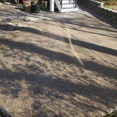 A paved patio area with scattered shadows, stairs, and planters beside a retaining wall in a backyard.