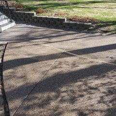 Curved concrete pathway with visible expansion joints, bordered by a stone retaining wall and grassy yard with trees.