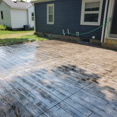 A stamped concrete patio with a wood plank pattern beside a house, partially wet from being recently washed or sealed.