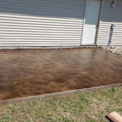 A newly installed brown stamped concrete patio adjacent to a house with beige siding and a white door.