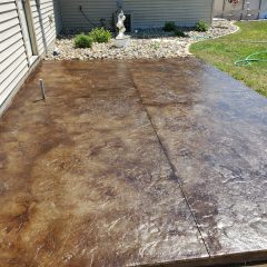 A stained concrete patio next to a house, bordered by landscaping rocks and a garden hose on nearby grass.