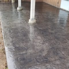 Wet stamped concrete patio with supporting columns, next to a brown house with windows and a white door.