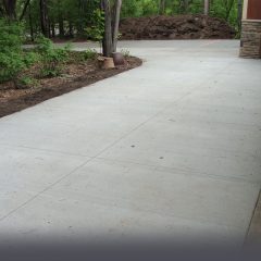 A newly paved concrete driveway curves through a wooded area with trees and shrubs on both sides.