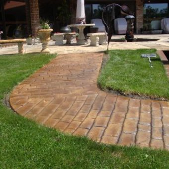 A stamped concrete walkway curves through a green lawn toward a patio with outdoor furniture and planters.