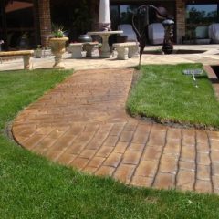 A stamped concrete walkway curves through a green lawn toward a patio with outdoor furniture and planters.