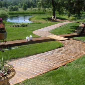 A winding brick pathway leads through a green lawn toward a pond, with potted plants and cleaning supplies nearby.
