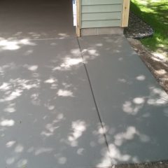 Freshly poured concrete driveway leading into a garage with light green siding, surrounded by grass and trees.
