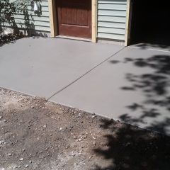 Freshly poured concrete patio in front of a door on a house with light green siding and wooden trim.