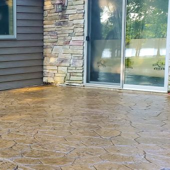 Stamped concrete patio with a stone and wood exterior wall, next to a glass sliding door reflecting trees and water.
