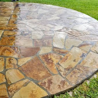 Circular stone patio with various shades of tan and brown, bordered by green grass and wet from recent rainfall.