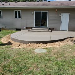 Freshly poured concrete patio with rounded edge behind a house, surrounded by dirt and grass.