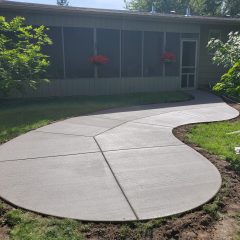 Freshly poured curved concrete walkway leading to a house with screened porch and red flowers in window boxes.